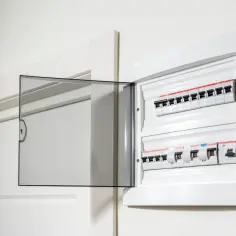 Otwarta biała skrzynka z bezpiecznikami elektrycznymi i przezroczystą klapką.